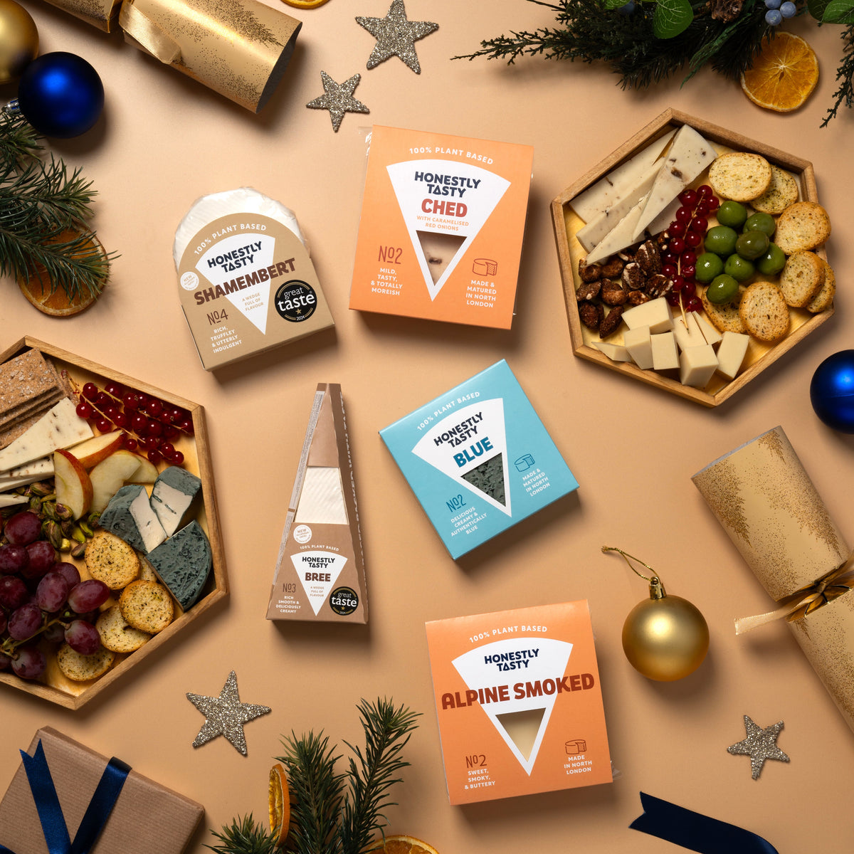 An overhead flat-lay of several packaged cheeses and charcuterie boards arranged with holiday decorations on a beige surface. Visible products include a round cheese labeled 'Shamembert', a wedge labeled 'Bree', a blue box labeled 'Blue', and orange boxes labeled 'Alpine Smoked'.