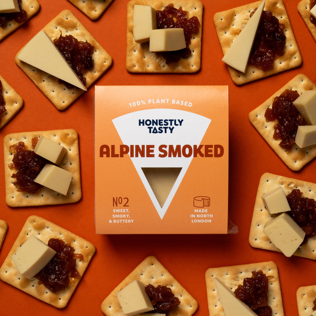A package of Honestly TastY Alpine Smoked crackers sits centered on a bright orange background, surrounded by multiple crackers topped with cheese wedges and dark red jam. The package is orange with white and dark red text.