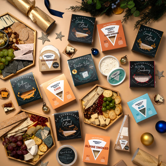 An overhead flat-lay of multiple gourmet cheese and charcuterie gift boxes arranged on a beige surface with holiday decorations, including pine branches, ornaments, stars, and a cracker. The boxes feature dark blue, orange, and light blue packaging with product names such as Alpine Smoked