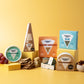 A styled product display of multiple artisan cheeses from Honeywell Farms on a solid yellow background. The cheeses include a round Garlic & Herb, a wedge of Alpine Smoked, a blue box of Blue, a wedge of Wed, and a box of Shamendment, accompanied by crackers, grapes, and a small bowl of nuts.