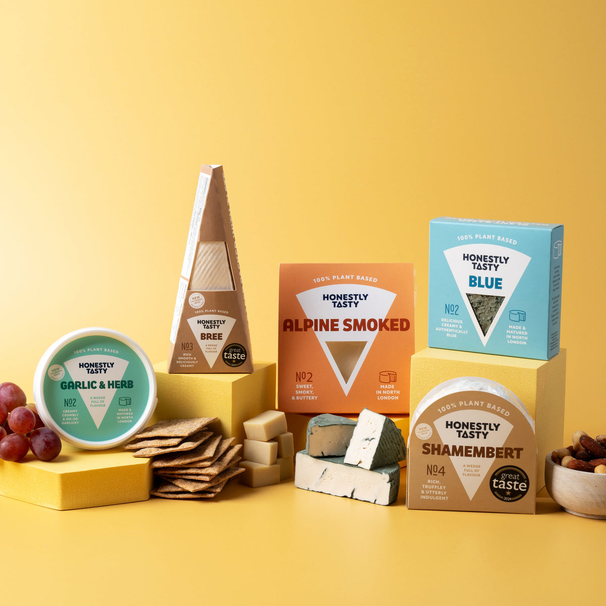 A styled product display of multiple artisan cheeses from Honeywell Farms on a solid yellow background. The cheeses include a round Garlic & Herb, a wedge of Alpine Smoked, a blue box of Blue, a wedge of Wed, and a box of Shamendment, accompanied by crackers, grapes, and a small bowl of nuts.