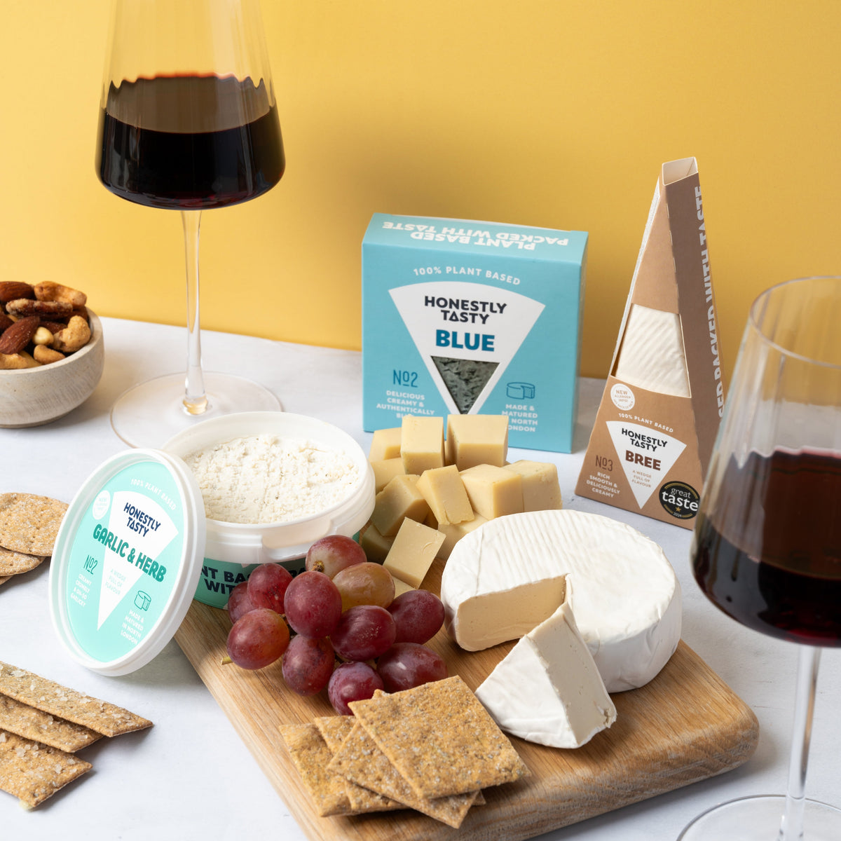 A styled cheese and wine charcuterie board setup on a white marble surface against a solid yellow background. The board contains various cheeses (including a wheel, wedges, and a block in packaging), crackers, red grapes, and a small bowl of nuts.