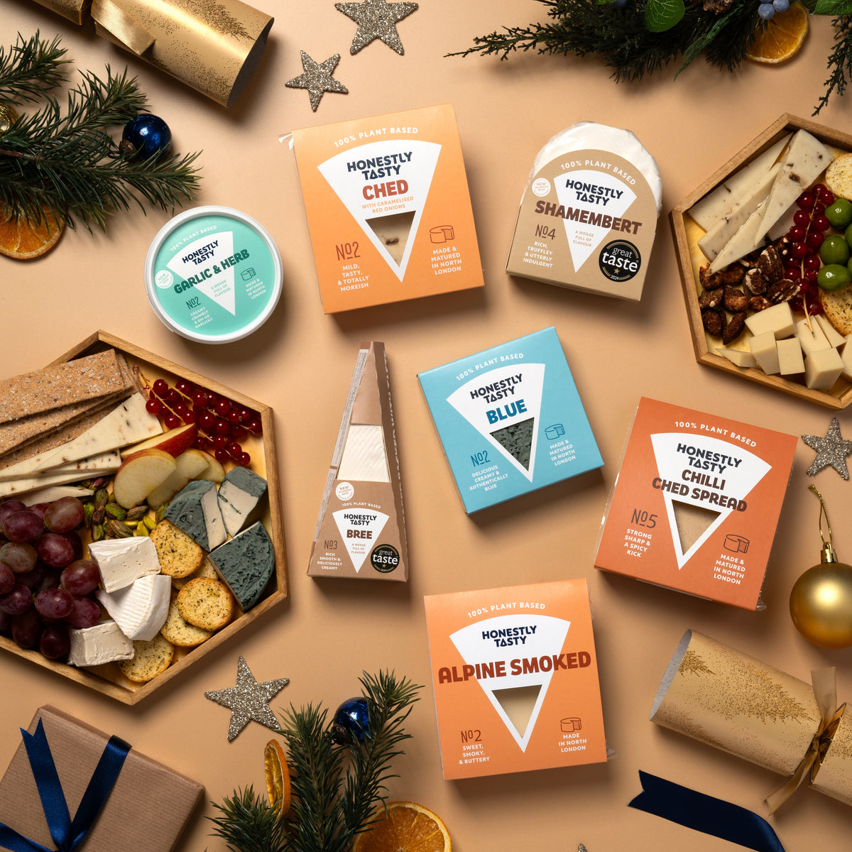 An overhead flat-lay of multiple boxes and boards of Monmouths Cheese & Charcuterie boards arranged on a beige surface with holiday decorations including pine branches, silver stars, a gold cracker, and wrapped gifts.