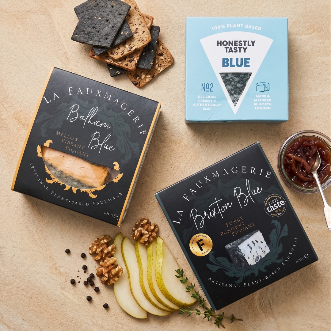Two boxes of 'La Fauxmagerie' plant-based blue cheese on a wooden surface with crackers, pears, and jam.