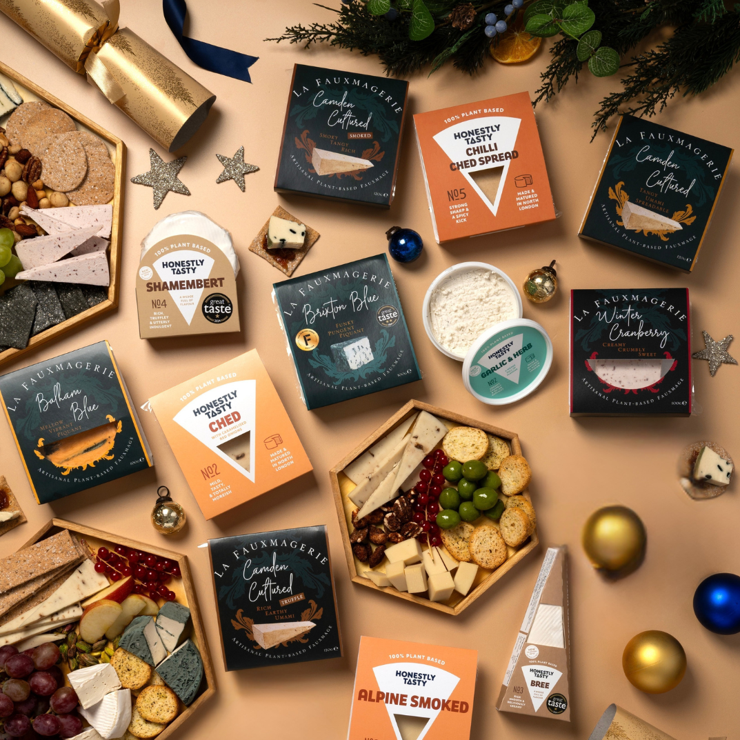 An overhead flat-lay of gourmet cheese and charcuterie boards arranged on a beige surface with holiday decorations. Multiple boxed and tray-style cheese spreads from Honestly Tasty Cheese Company are visible, including smoked and brie varieties, alongside crackers, grapes, nuts, gold and blue ornaments, wrapped gifts, and festive greenery.